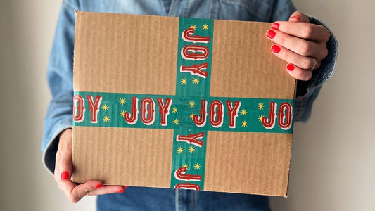 A woman holiday a gift box sealed with cute red and green packing tape with Joy printed on it.