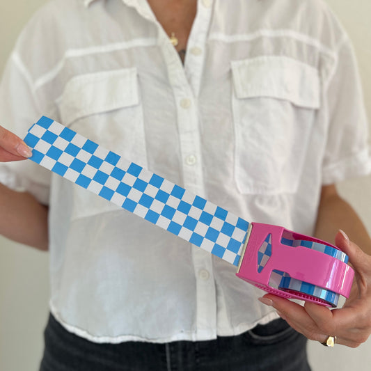 Woman holding a mini roll of blue checkered packing tape with the tape pulled out from a pink dispenser to show the printed pattern.