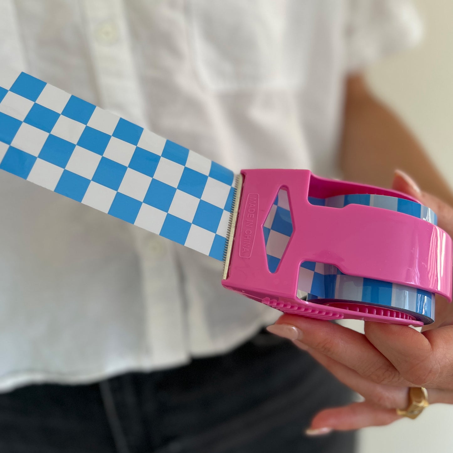 Woman holding a mini roll of blue checkered packing tape with the tape pulled out from a pink dispenser to show the printed pattern.
