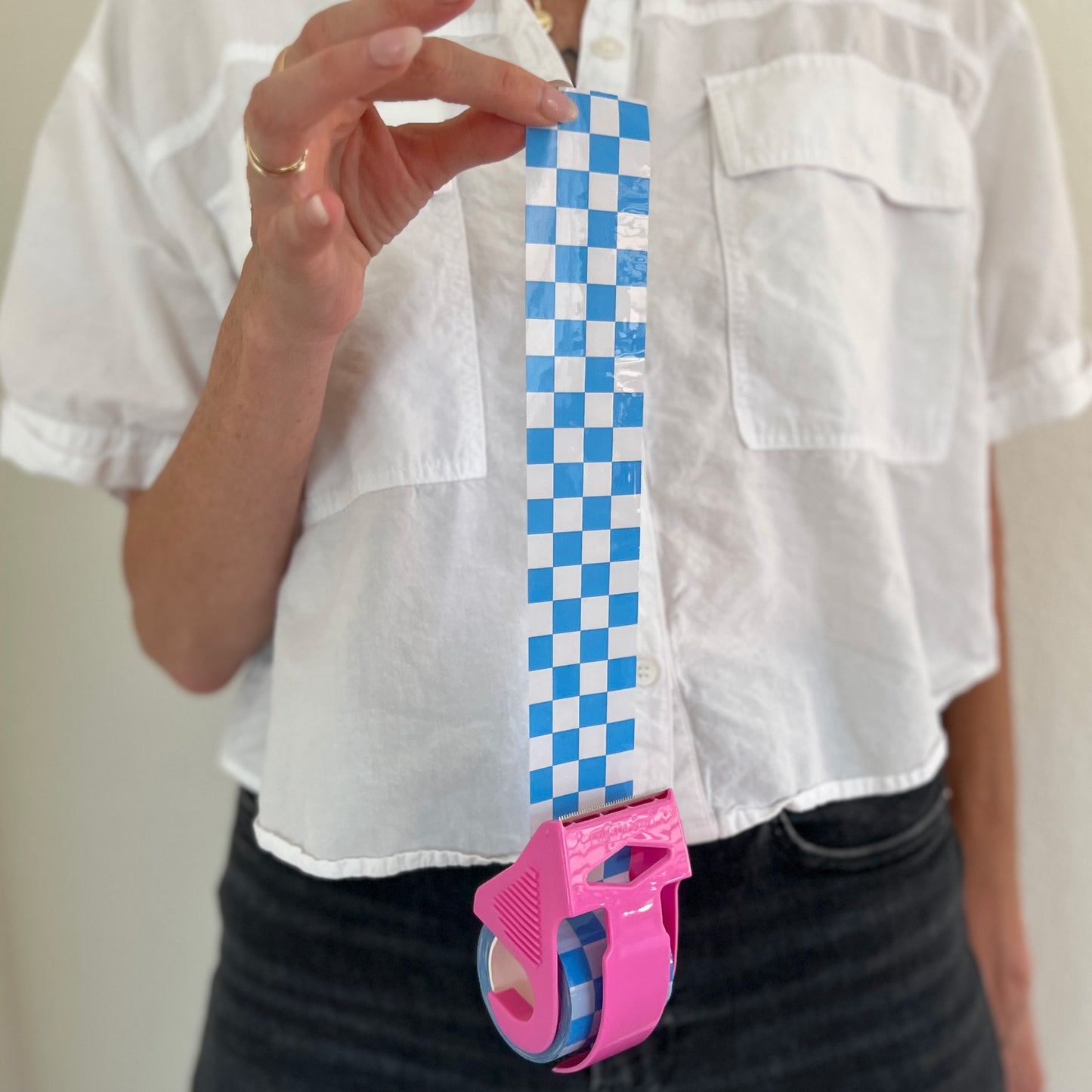 Woman holding a mini roll of blue checkered packing tape with the tape pulled out from a pink dispenser to show the printed pattern.