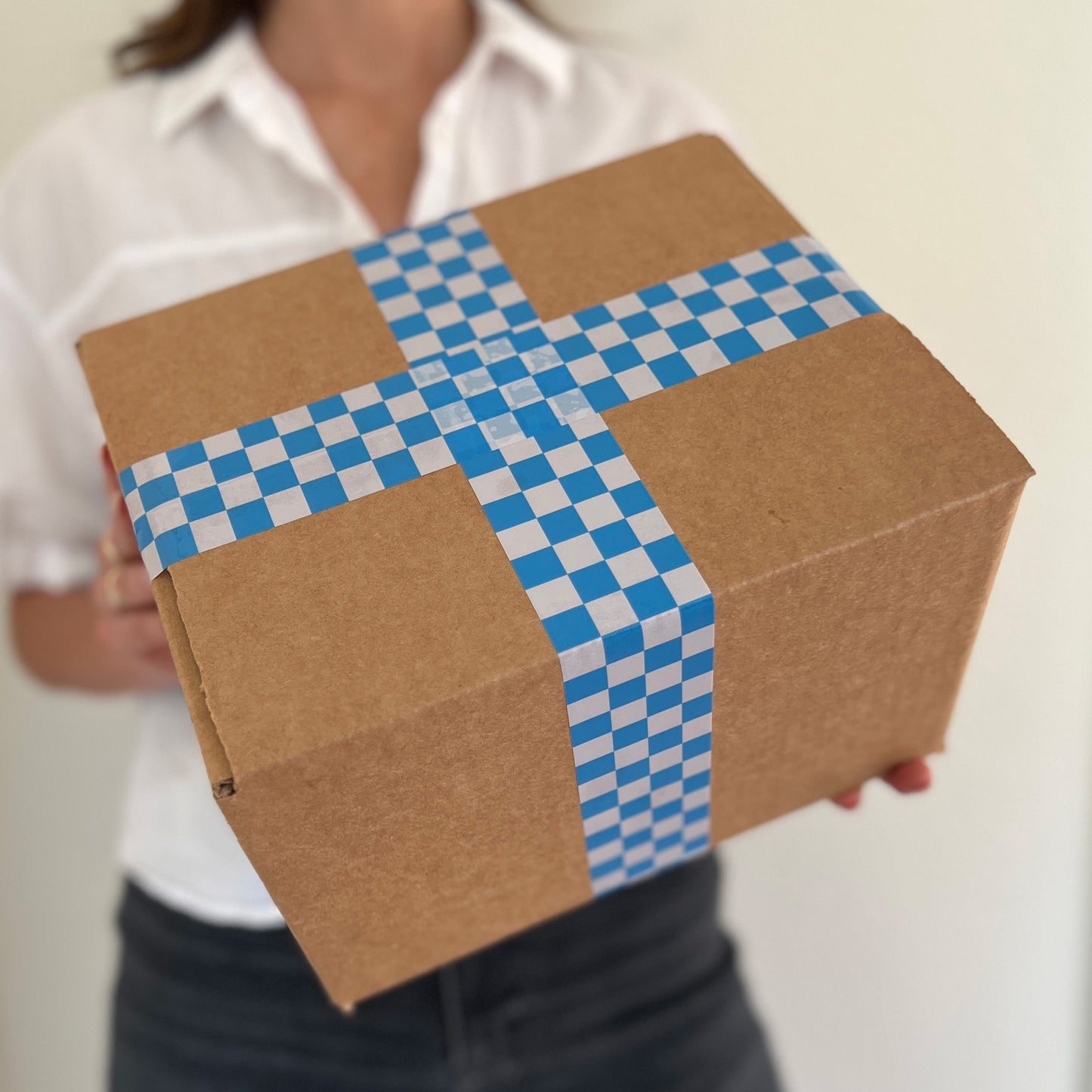 Woman holding a cardboard box sealed with blue checkered packing tape and a pink dispenser.
