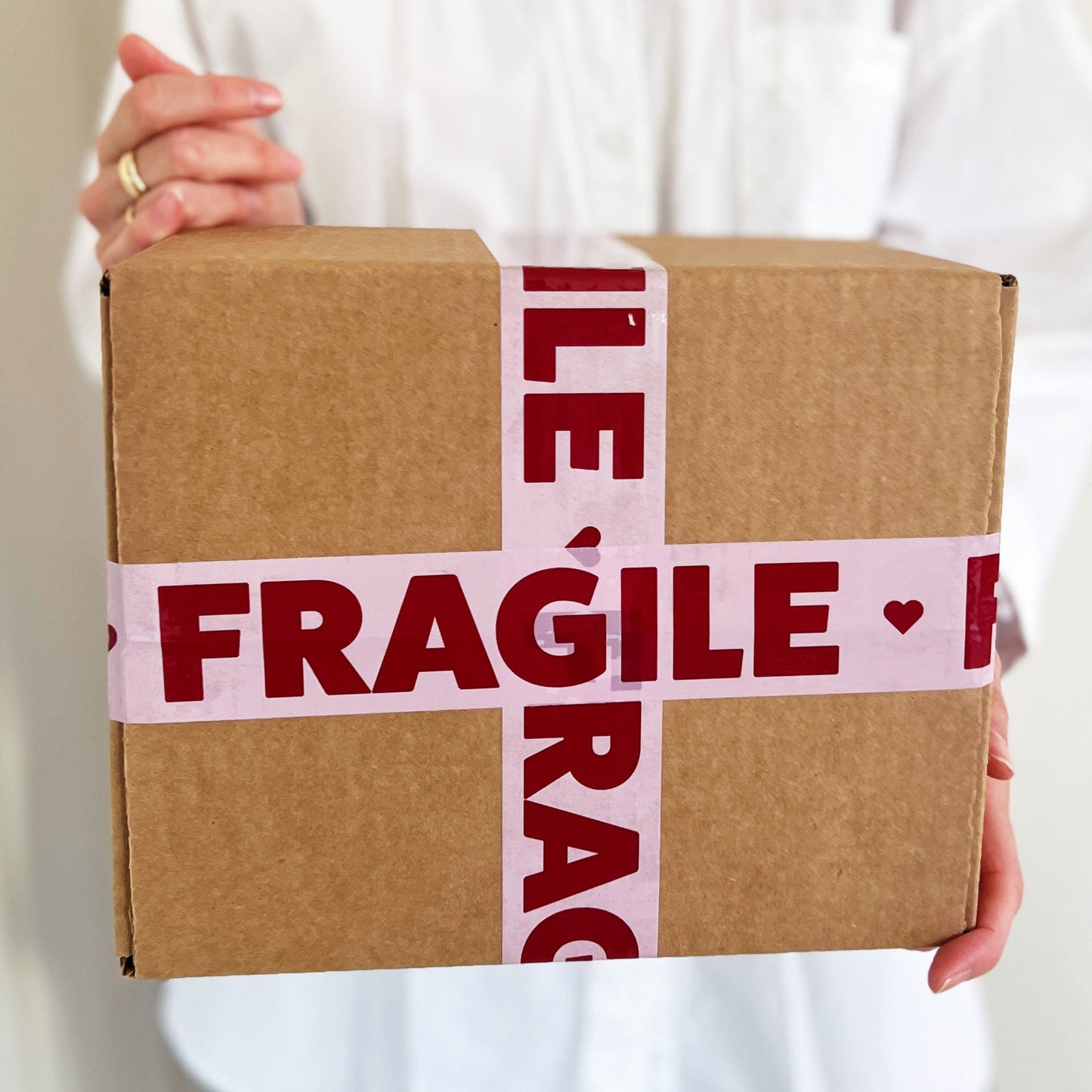 A woman holding a cardboard box sealed with pink packing tape that reads “Fragile” in bold burgundy text—perfect for protecting breakable items during shipping or moving.