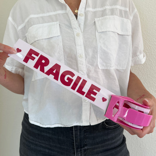 Woman holding a roll of pink packing tape with burgundy “Fragile ♥️” text pulled out to show the print, in a matching pink dispenser—ideal for sealing moving boxes or shipping delicate e-commerce orders with extra care.