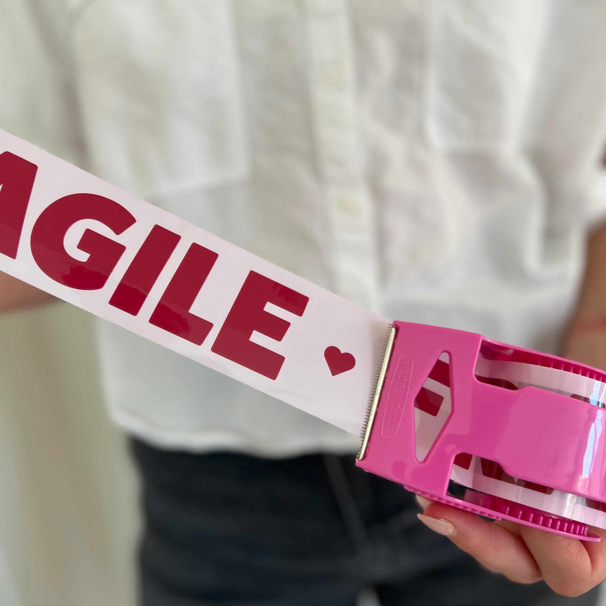 Woman holding a roll of pink packing tape with burgundy “Fragile” text pulled out to show the print, in a matching pink dispenser—ideal for sealing moving boxes or shipping delicate e-commerce orders with extra care.