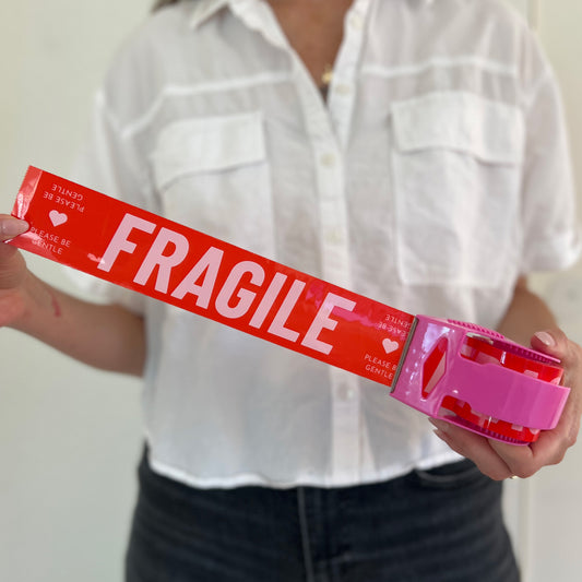 Woman holding red packing tape with pink Fragile Please Be Gentle text in a pink dispenser for shipping delicate items.