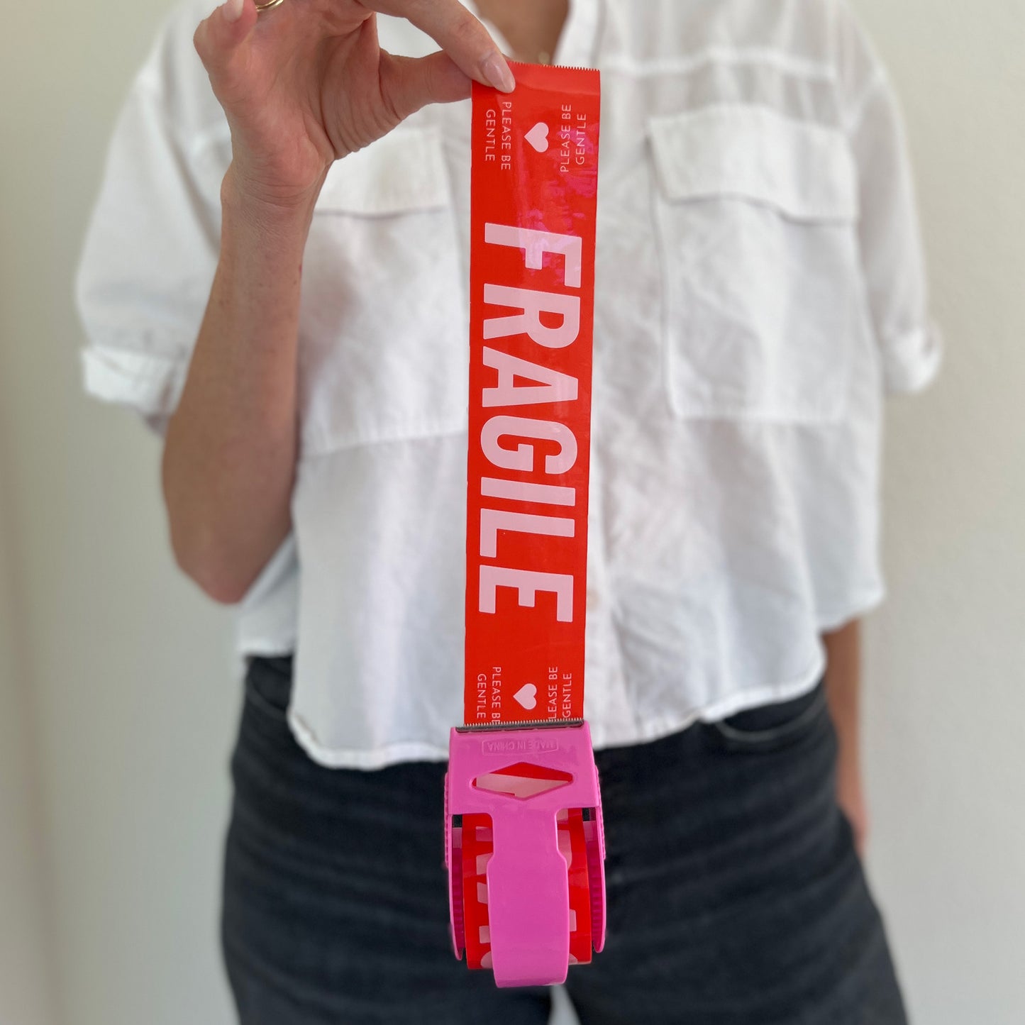 Woman holding red packing tape with pink Fragile Please Be Gentle text in a pink dispenser for shipping delicate items.