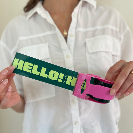 Woman holding out a roll of green packing tape with bold mint “Hello! I’m Here” text in a pink dispenser—designed to add fun, personality, and visibility to small business shipments.