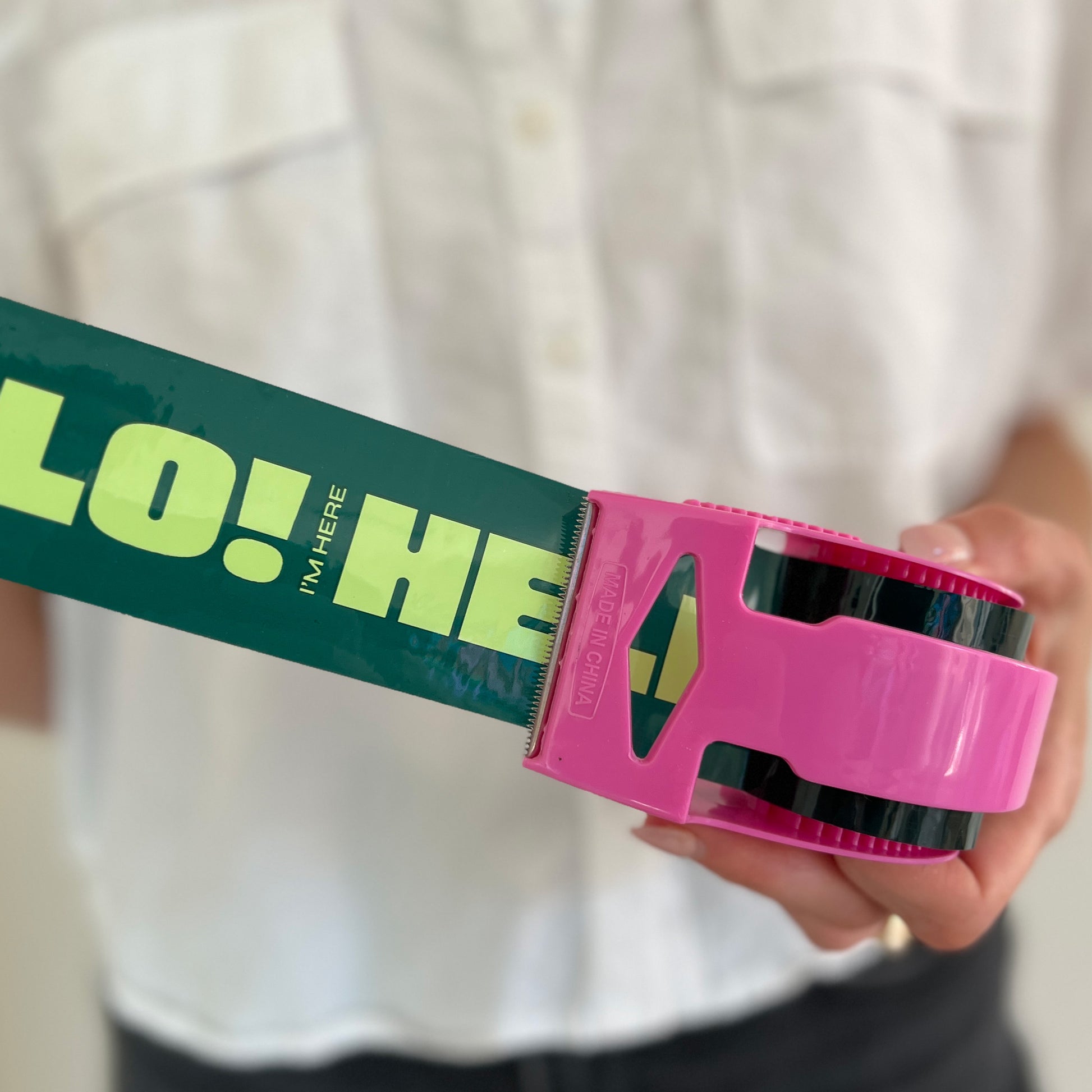 Woman holding out a roll of green packing tape with bold mint “Hello! I’m Here” text in a pink dispenser—designed to add fun, personality, and visibility to small business shipments.