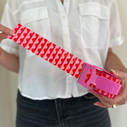 Woman holding a mini roll of pink and red wave packing tape with the tape pulled out from the dispenser to show the bold, colorful design—perfect for decorative shipping, gift packaging, and small business orders.
