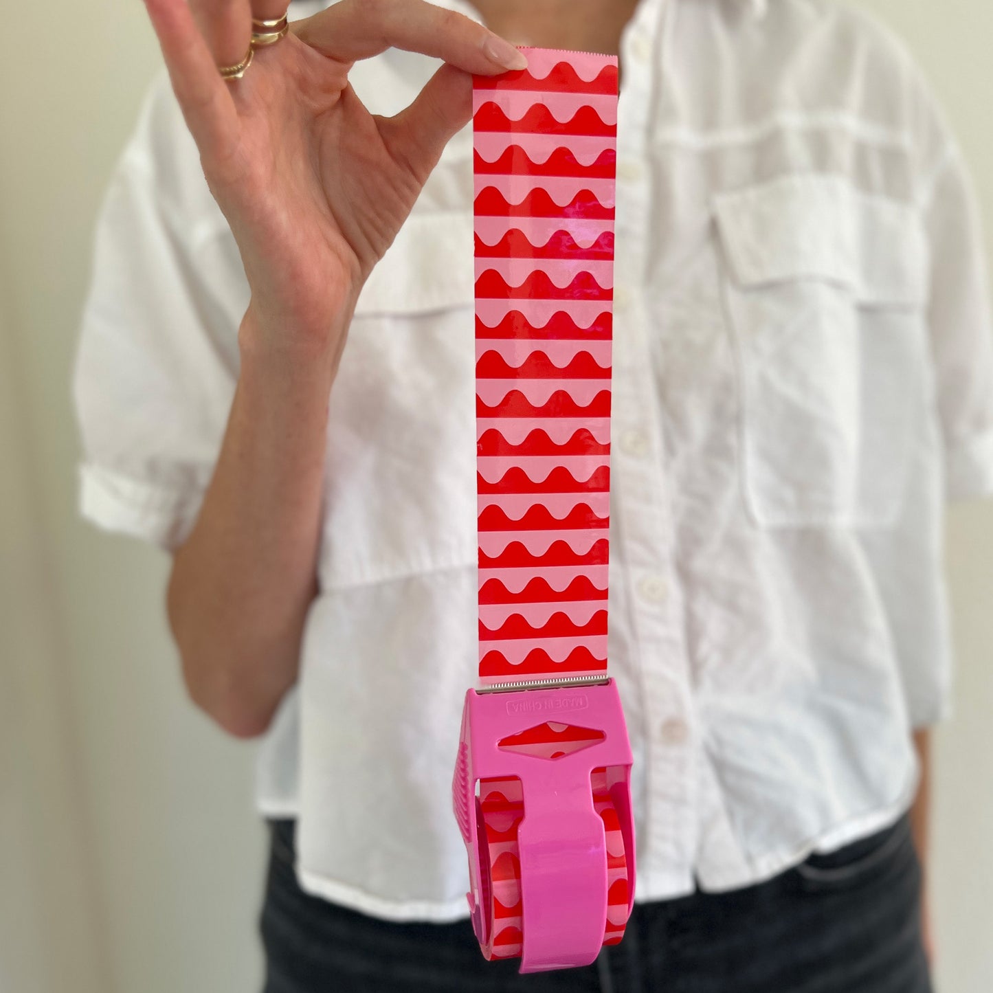 Woman holding a mini roll of pink and red wave packing tape with the tape pulled out from the dispenser to show the bold, colorful design—perfect for decorative shipping, gift packaging, and small business orders.