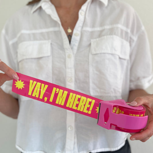 Woman holding a roll of pink packing tape with bold yellow “Yay, I’m Here!” text in a bright pink dispenser—designed to add fun, personality, and professionalism to small business packaging.