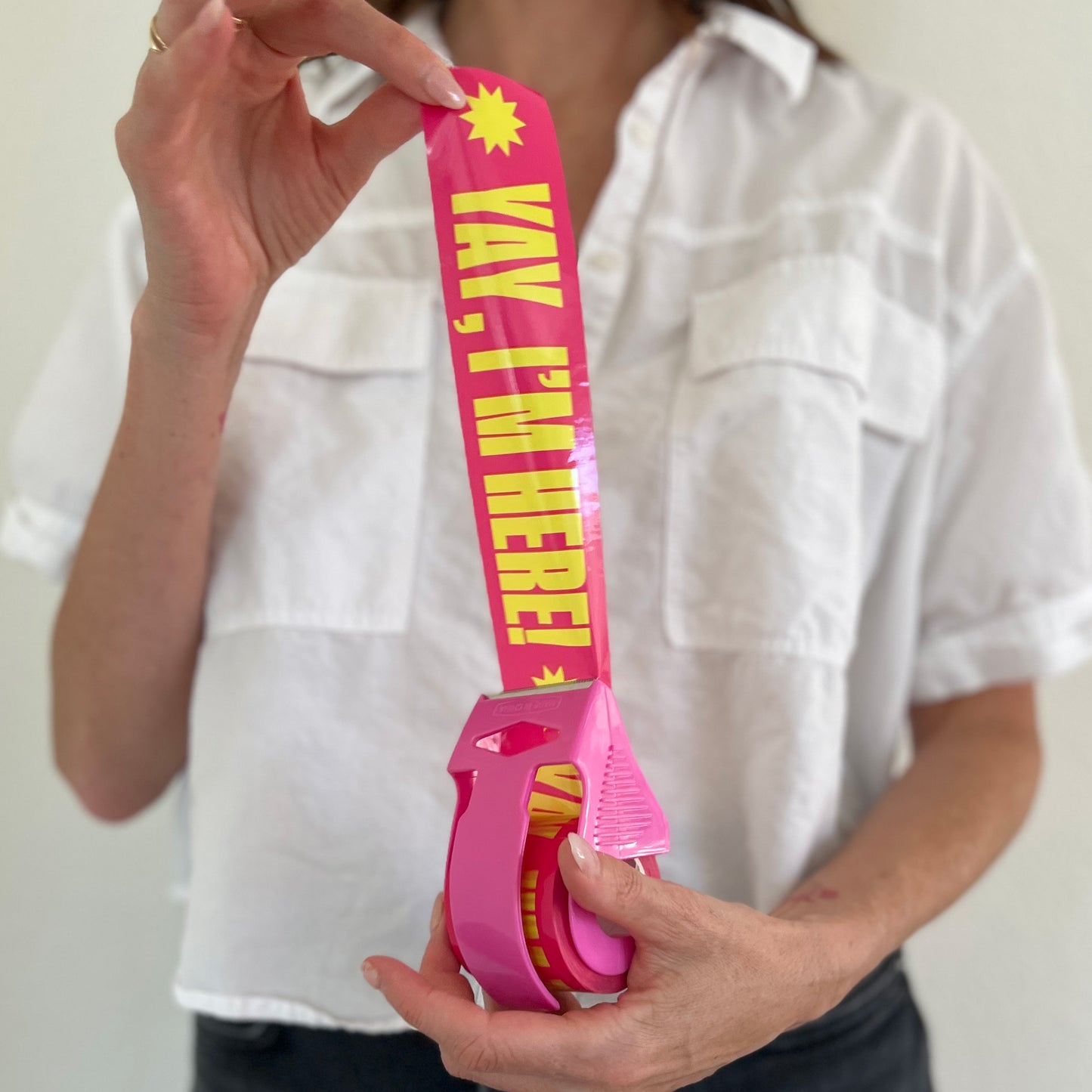 Woman holding a roll of pink packing tape with bold yellow “Yay, I’m Here!” text in a bright pink dispenser—designed to add fun, personality, and professionalism to small business packaging.