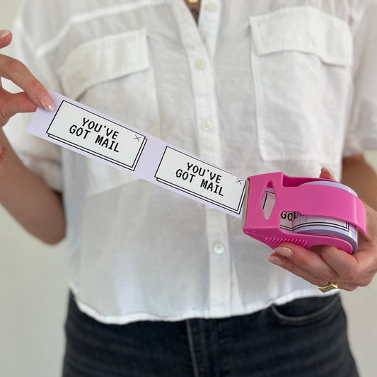 Woman holding a mini roll of “You’ve Got Mail” printed packing tape in a bright pink dispenser; nostalgic 90s-style design with black text on white tape, perfect for small business packaging.