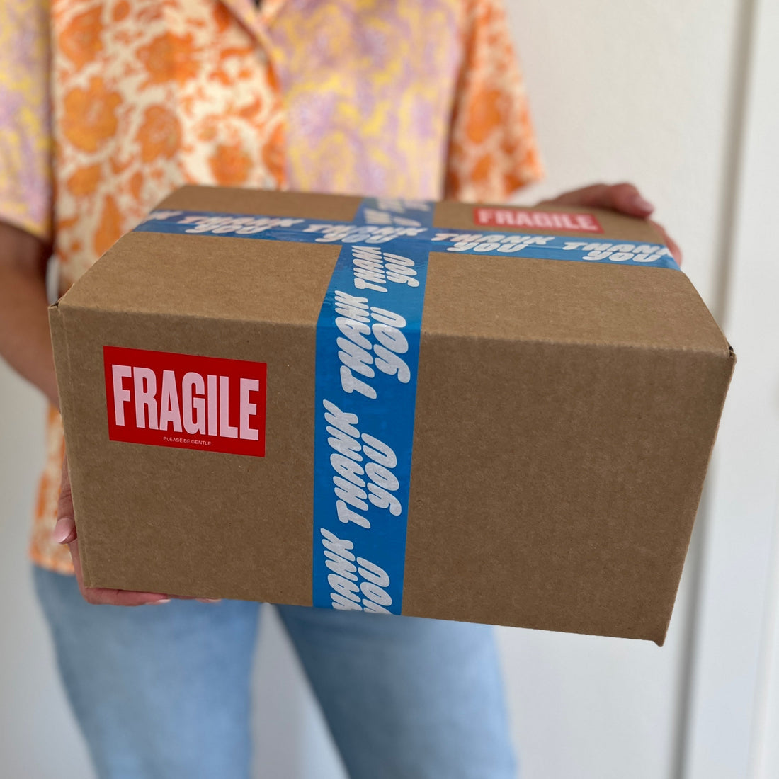 Woman holding a standard brown shipping box sealed with colorful “Thank You” tape and a Fragile sticker—example of USPS-compliant packaging for Poshmark Ground Advantage.