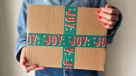 A woman holiday a gift box sealed with cute red and green packing tape with Joy printed on it.