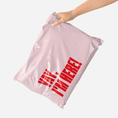 Pink shipping envelope with red text that reads "Yay, I'm Here!" held by a hand on a white background