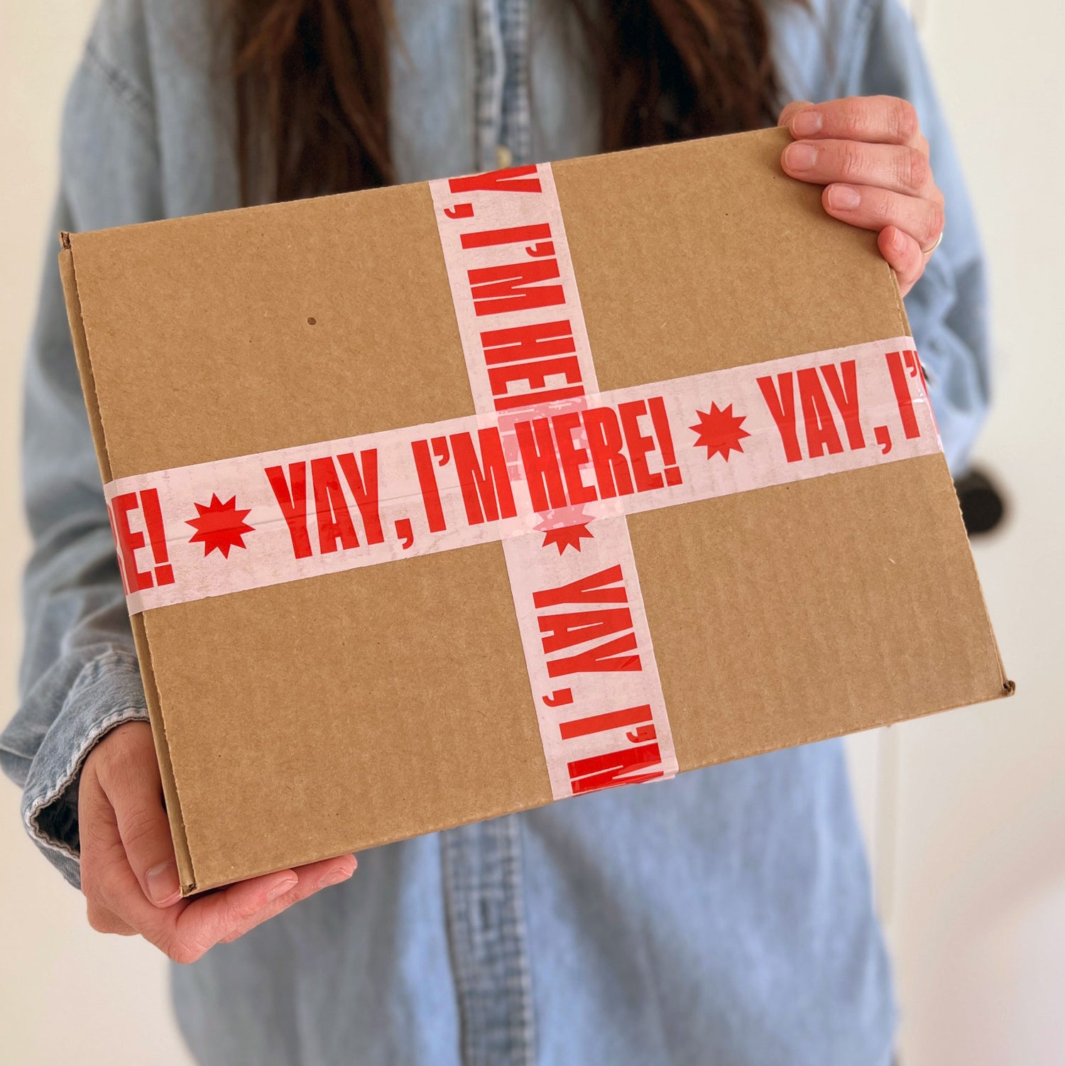 Person holding a cardboard box with 'YAY, I'M HERE!' tape
